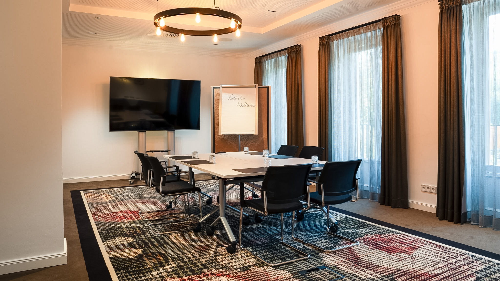 Tagungsraum Gutsstube auf Gut Altona - Blocktisch mit acht Stühlen, Monitor und Flipchart vor bodentiefen Fenstern